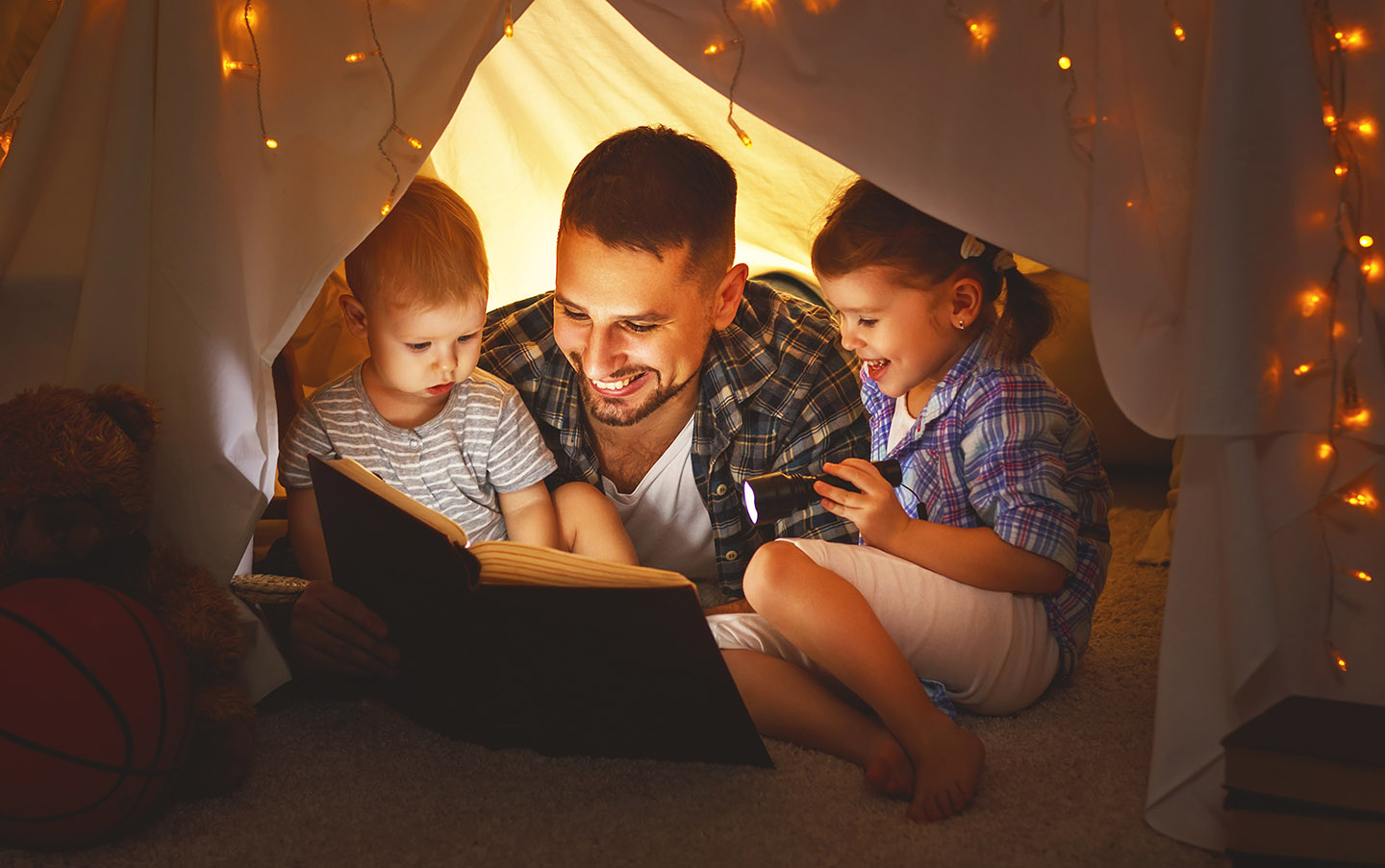 Father and two children reading in a blanket fort with twinkling lights