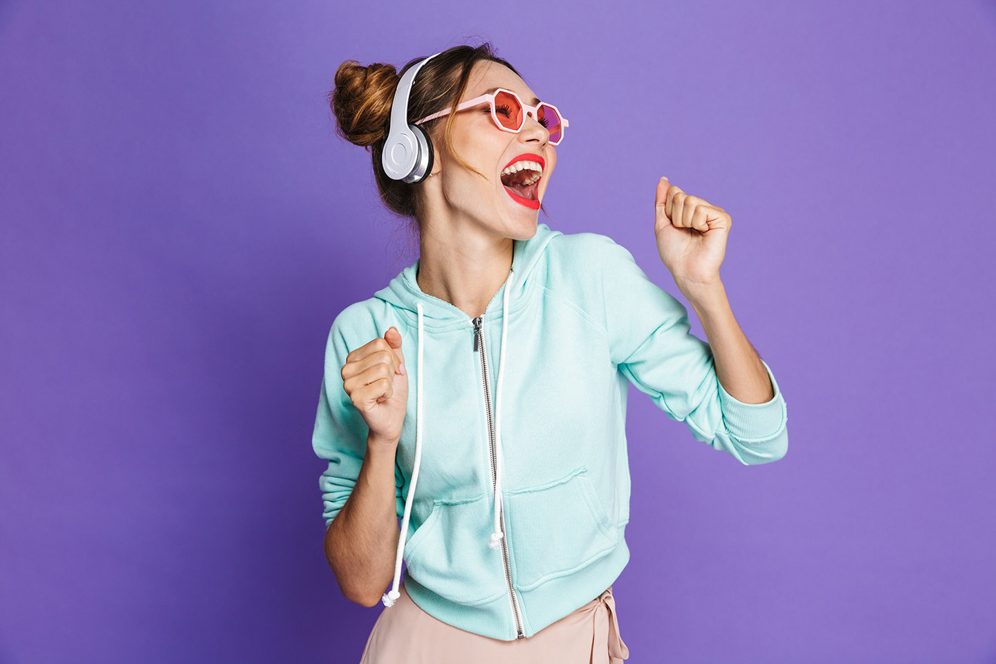 Portrait of a happy young girl with bright makeup over violet background, listening to music with headphones