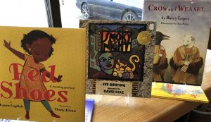 Children's books on a shelf in front of a window including Red Shoes and Crow and Weasel