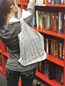 A woman carries a reusable shopping bag over one shoulder, the bag has flower print, and fills it with books from an orange bookshelf. Earth Day is a great day to update your totes.