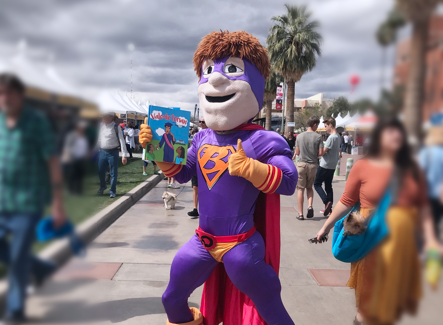 Bookmans Book Man mascot in a purple and orange costume with a cape holding a children's book at the 2019 Tucson Festival of Books